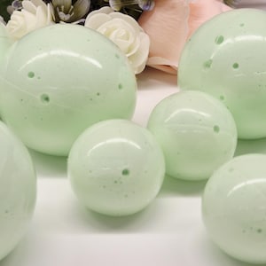 May include: Several light green, spherical bath bombs of varying sizes are arranged in a group. The bath bombs have a smooth, slightly textured surface with small holes. A bouquet of pink and white flowers is in the background.