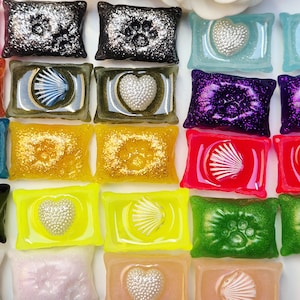 Könnte beinhalten: Eine Sammlung farbenfroher Dekorationsstücke aus Kunstharz in verschiedenen Formen. Einige Stücke weisen Glitzer, Pfotenabdrücke, Herzen und Muschelmotive auf. Farben sind Schwarz, Blau, Lila, Gelb, Rot, Grün und Pfirsich.