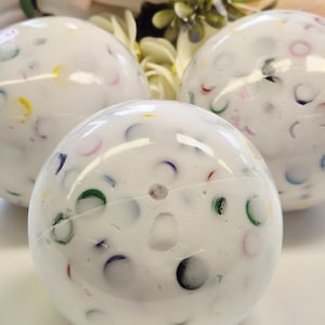 Peut inclure: Trois objets décoratifs sphériques blancs avec des accents colorés en demi-cercle. Les sphères ont un aspect moucheté avec diverses couleurs, dont le rouge, le jaune, le vert et le bleu. Le fond présente des fleurs douces et floues.