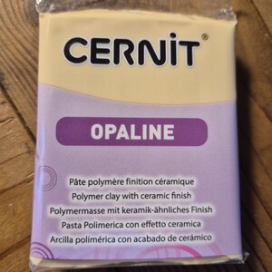 Może przedstawiać: Blok jasnożółtej gliny polimerowej z ceramicznym wykończeniem. Opakowanie zawiera nazwę marki "CERNIT" i nazwę koloru "OPALINE" w kolorze fioletowym. Tekst na opakowaniu brzmi "Pâte polymère finition céramique, Polymer clay with ceramic finish, Polymermasse mit keramik-ähnliches Finish, Pasta Polimerica con effetto ceramica, Arcilla polimérica con acabado de cerámico."