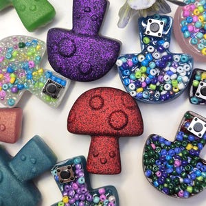 Könnte beinhalten: Eine Sammlung farbenfroher, glitzernder Resin-Anhänger in verschiedenen Formen, darunter Pilze und abstrakte Formen. Einige Anhänger sind mit kleinen, bunten Perlen gefüllt und haben eine kleine, quadratische Metallverzierung. Die Farbpalette umfasst Rot, Lila, Grün und Blau.