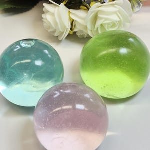 Peut inclure: Trois sphères de verre transparentes, dans les tons de turquoise, vert et rose. Les sphères sont lisses et rondes, reflétant la lumière. Des fleurs artificielles blanches et roses sont en arrière-plan, suggérant un thème décoratif.