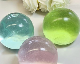 Puede incluir: Tres esferas de vidrio transparente en tonos turquesa, verde y rosa. Las esferas son lisas y redondas, reflejando la luz. Flores artificiales blancas y rosas en el fondo sugieren un tema decorativo.
