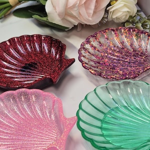 Può includere: Quattro portagioie a forma di conchiglia nei toni del rosso, viola, rosa e verde. Ogni piatto è impreziosito da glitter, che aggiungono un effetto scintillante. I piatti sono disposti su una superficie bianca, con decorazioni floreali sullo sfondo.