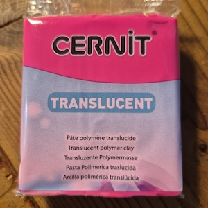 May include: A pink package of translucent polymer clay by the brand Cernit. The package has the text "TRANSLUCENT" in white letters on a blue background. The text "Pâte polymère translucide, Translucent polymer clay, Transluzente Polymermasse, Pasta Polimerica traslucida, Arcilla polimérica translúcida" is printed in black on a white background.