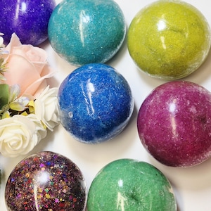 Könnte beinhalten: Eine Sammlung von sieben kugelförmigen Badebomben in verschiedenen Farben, darunter Violett, Türkis, Limettengrün, Blau, Magenta und eine dunkle, glitzernde, mehrfarbige Kugel, angeordnet neben einem Bündel aus blassrosa und weißen künstlichen Rosen.