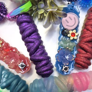 Pode incluir: Uma variedade de acessórios coloridos feitos à mão em resina e cordão. A imagem apresenta um cordão roxo brilhante, uma peça de resina azul com um rosto sorridente e outras peças com enfeites florais e geométricos. Os itens estão dispostos em uma superfície branca.