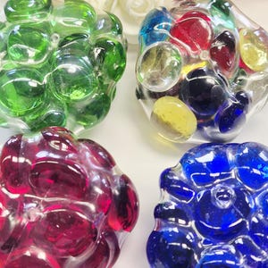 Può includere: Quattro grappoli decorativi in vetro, ciascuno riempito di biglie di vetro colorate. I grappoli sono nelle tonalità di verde, rosso, blu e un mix di colori. Le biglie sono rotonde e lisce, creando un'esposizione giocosa e vibrante.