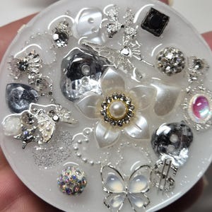 Pode incluir: Um suporte de telefone redondo branco com uma variedade de elementos decorativos, incluindo uma flor branca com um centro de pérola, uma borboleta e strass.