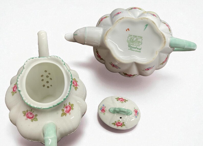 May include: A white porcelain teapot set with a floral rosebud pattern. The set includes the teapot, lid, and a strainer. The teapot has a scalloped edge and a mint green handle and spout. The underside of the teapot has the text "Shelley England Rosebud".