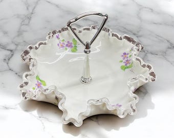 Puede incluir: Una fuente blanca de dos niveles con asa plateada. El plato tiene un borde con volantes con ribete plateado y está decorado con detalles florales morados. Diseñado para servir postres o aperitivos.