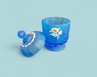 May include: A blue glass trinket box with a lid, featuring a white and yellow daisy design. The box is open, revealing the interior. The lid has a decorative edge. The box sits on a light blue background.