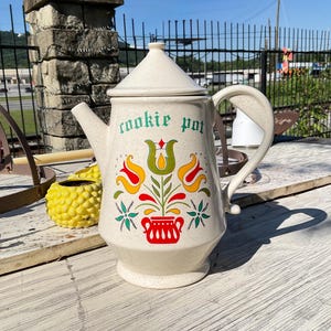 Op de afbeelding: Een crèmekleurige keramische koekjespot met een conisch deksel en een handvat. De voorzijde is voorzien van de tekst "cookie pot" in groene letters boven een kleurrijk bloemmotief met rode, gele en groene tulpen en een rode onderkant.