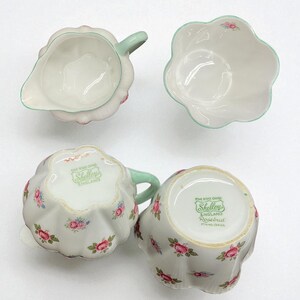 May include: A vintage porcelain tea set featuring a creamer, sugar bowl, and two teacups. The set is white with pink rose floral patterns and mint green accents. The bottom of the teacups and creamer have the text "Shelley England".