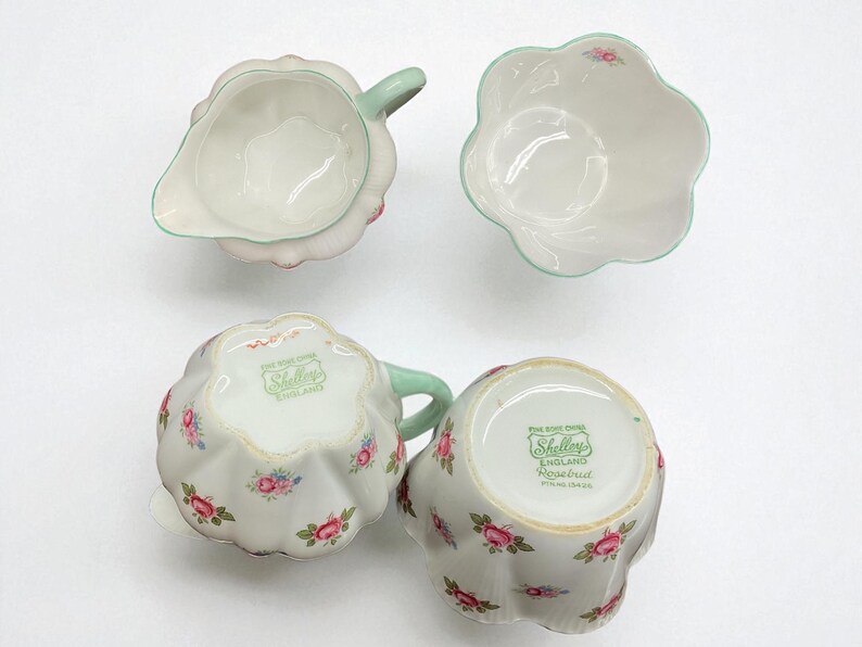 May include: A vintage porcelain tea set featuring a creamer, sugar bowl, and two teacups. The set is white with pink rose floral patterns and mint green accents. The bottom of the teacups and creamer have the text "Shelley England".