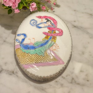 May include: A white oval trinket box with a silver trim. The lid features a hand-painted design of a woman in a pink and blue robe with a green and gold background.