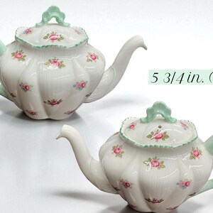 May include: White ceramic teapot with a floral pattern of pink roses and green accents. The handle and lid handle are light green. The teapot is 5 3/4 inches (14.6 cm) tall.