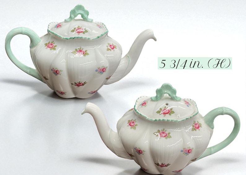 May include: White ceramic teapot with a floral pattern of pink roses and green accents. The handle and lid handle are light green. The teapot is 5 3/4 inches (14.6 cm) tall.