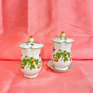 May include: Two white ceramic jars with lids, decorated with a strawberry motif. The jars have a bulbous shape and are adorned with green leaves and red and yellow strawberries. The lids are topped with a small, orange-tipped knob.