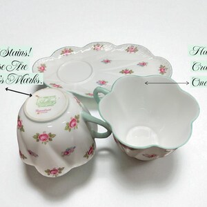 May include: A vintage teacup set featuring a cup, saucer, and plate. The white porcelain pieces are adorned with pink roses and green accents. The cup has a hairline crack. Text on the cup reads "Not Stains! These Are Painter's Marks."