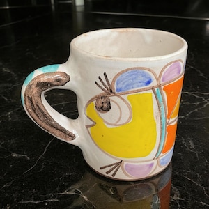 May include: White ceramic mug with a hand-painted design of a yellow fish with blue, orange, and purple accents. The mug has a brown handle.