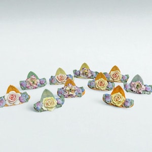 Pode incluir: Uma coleção de chapéus florais em miniatura. Cada chapéu apresenta uma rosa central, rodeada por flores menores em tons de rosa, amarelo e roxo. Os chapéus têm abas douradas ou verdes.