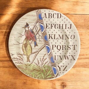 May include: A decorative ceramic plate with a bird illustration in blue, red, and yellow. The plate features the alphabet and a decorative blue and brown border. The plate is a decorative item.