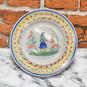 Op de afbeelding: Een wit keramiek bord met een blauwe, gele en rode rand. Het midden van het bord heeft een handgeschilderd tafereel van een persoon in een groen jasje en een blauwe rok die in een bloemenveld staat.
