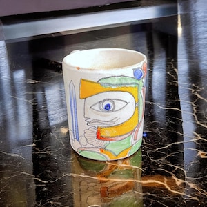May include: A white ceramic mug with a colorful abstract design featuring a large eye with a blue iris. The design includes yellow, green, red, and blue colors.