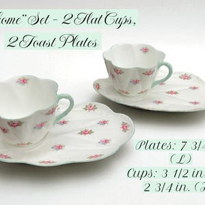 May include: A floral-patterned teacup and saucer set. The set includes two cups and two plates. The plates are 7.75 inches long, and the cups are 3.5 inches wide and 2.75 inches high. The set is decorated with pink roses and a mint green trim.