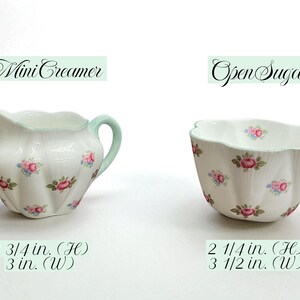 May include: A white porcelain mini creamer and open sugar bowl set. The creamer has a mint green handle and rim, and the sugar bowl has a scalloped edge. Both are decorated with pink and blue rose clusters. The creamer is 2 3/4 inches high and 3 inches wide. The sugar bowl is 2 1/4 inches high and 3 1/2 inches wide.