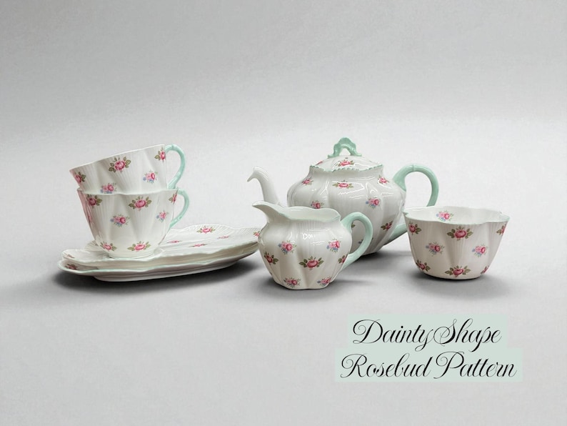 May include: A white porcelain tea set with a rosebud pattern, including teacups, saucers, a teapot, creamer, and a sugar bowl. The handles and accents are a light teal color. The text "Dainty Shape Rosebud Pattern" is visible.