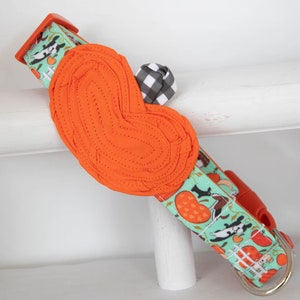 May include: A teal and orange dog collar with a heart-shaped orange buckle and a black and white checkered bow. The collar features a pattern of dogs and pumpkins.