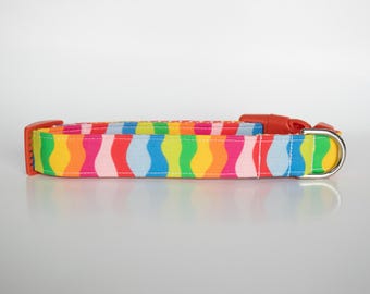 Puede incluir: Un collar de perro colorido con un patrón de ondas arcoíris. El collar está hecho de tela y tiene una hebilla de plástico roja y un anillo en D de metal plateado.