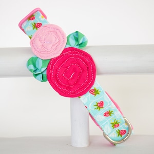 Puede incluir: Un collar para perro azul claro con estampado de fresas y un adorno de flor de tela rosa y roja.