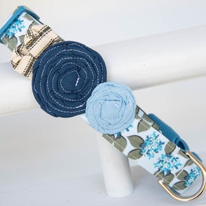 Puede incluir: Un collar de perro azul con un patrón floral y tres adornos de flores de tela en tonos azules. El collar tiene una hebilla de metal dorado.
