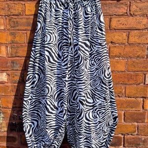 May include: Loose-fitting trousers featuring a black and white zebra stripe pattern. The garment has an elasticated waistband with a drawstring and gathered leg openings that include small ties at the bottom, hanging from a wooden clothes hanger.