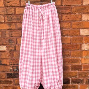 May include: Loose-fitting trousers featuring a pink and white gingham check pattern. The garment has an elasticated waistband with a matching fabric drawstring tie and gathered, adjustable tie-up cuffs at the bottom of each leg for a tapered look.