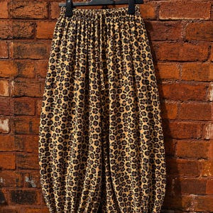 May include: Wide-leg trousers featuring a brown, black, and tan leopard print pattern. The garment has an elasticized waistband with a drawstring, a loose, voluminous silhouette, and adjustable tie-cuff hems at the bottom of each leg.