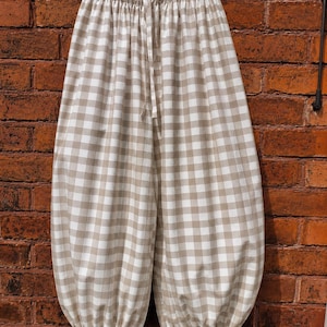 May include: Beige and white gingham patterned trousers featuring a loose, voluminous silhouette. The pants have an elasticized waistband with a drawstring tie and gathered leg openings that are finished with small, adjustable fabric ties at each hem.