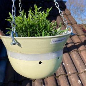 Pode incluir: Um vaso de plantas pendurado de cor verde claro feito de um capacete reutilizado com uma faixa branca no meio. O vaso está pendurado em uma corrente prateada e contém uma planta verde.