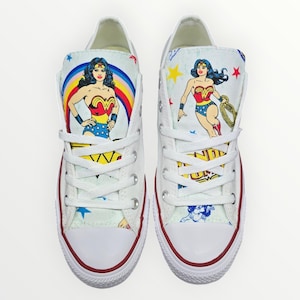 Puede incluir: Zapatillas bajas de lona blancas con un gráfico de Wonder Woman en los laterales. El gráfico presenta a Wonder Woman con su icónico traje rojo, azul y dorado. Las zapatillas tienen cordones blancos y un borde rojo alrededor de la suela.