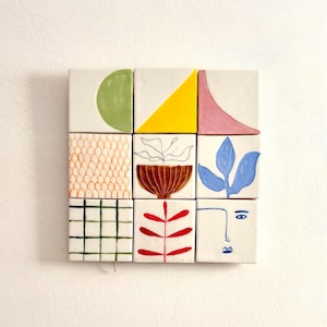 Peut inclure: Une œuvre d'art murale en céramique blanche avec un motif géométrique. Les carreaux sont disposés en grille et présentent diverses formes, couleurs et motifs, notamment un cercle vert, un triangle jaune, une courbe rose, un pot brun avec une plante, une feuille bleue, une branche rouge et un visage stylisé.