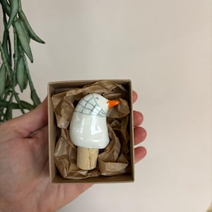 Könnte beinhalten: Ein Keramik-Flaschenverschluss in Form eines weißen Vogelkopfes mit leuchtend orangefarbenem Schnabel und einem dunkelgrünen Gittermuster auf dem Kopf. Der Verschluss hat einen Naturkorken und liegt in einer kleinen braunen Pappschachtel.