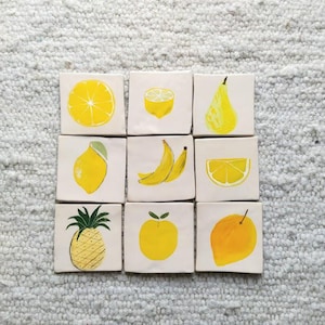 Puede incluir: Nueve azulejos de cerámica blanca con diseños de frutas pintadas en amarillo. Los azulejos presentan un limón, una pera, un plátano, una piña, una naranja y una rodaja de limón.