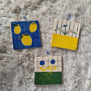 May include: Three ceramic tiles with different designs. The first tile has a blue background with three yellow lemons. The second tile has a yellow and white striped background with a blue flower. The third tile has a white background with a green and yellow flower with blue petals.