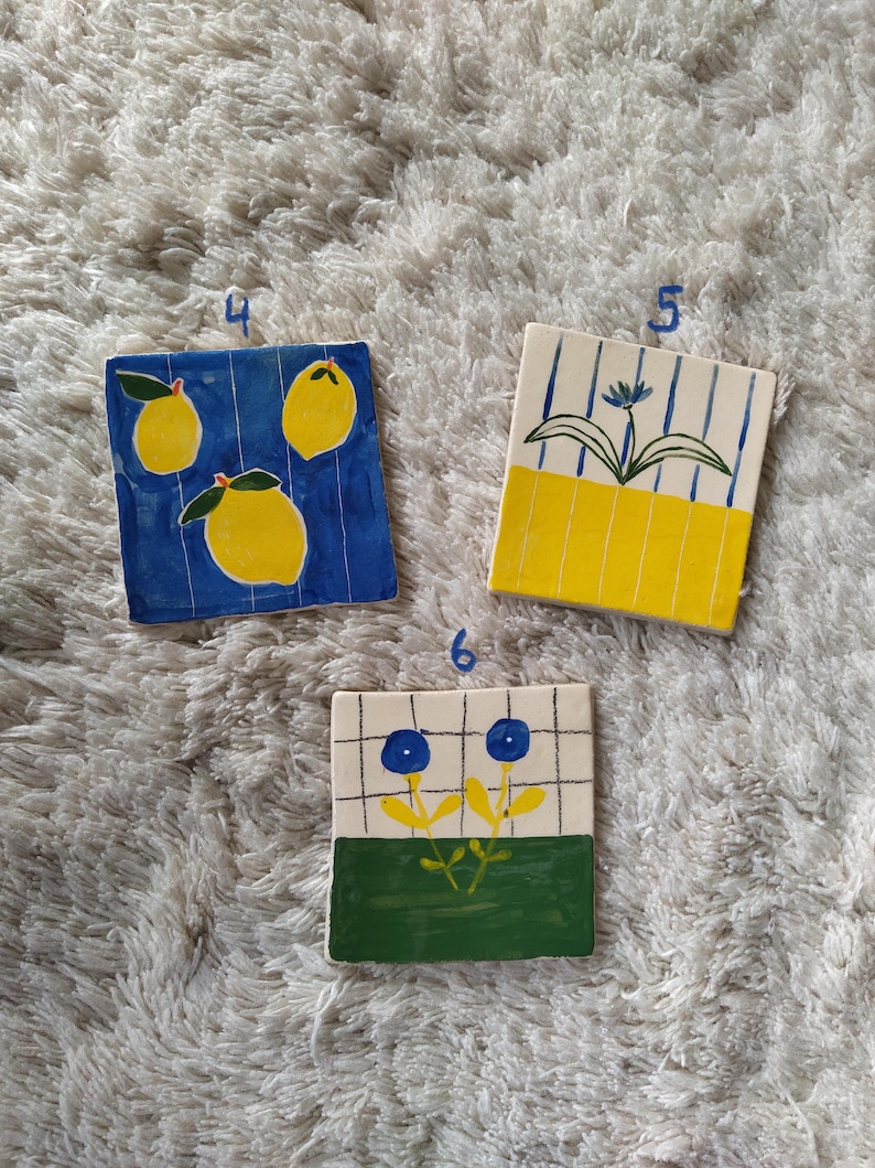 May include: Three ceramic tiles with different designs. The first tile has a blue background with three yellow lemons. The second tile has a yellow and white striped background with a blue flower. The third tile has a white background with a green and yellow flower with blue petals.