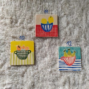 May include: Three ceramic tiles with hand-painted fruit designs. The first tile has a yellow background with a green basket of oranges. The second tile has a red and white checkered background with a blue bowl of lemons. The third tile has a blue and white striped background with a red and white striped bowl of pears.