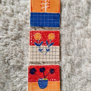 May include: Three square ceramic tiles with hand-painted floral designs. The top tile has a blue and orange design with a white flower. The middle tile has a red background with two yellow flowers. The bottom tile has a red background with three black flowers in a blue vase.