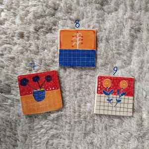 May include: Three square ceramic tiles with different floral designs. The first tile has a red background with black flowers in a blue vase. The second tile has an orange background with a white tree and blue grid. The third tile has a red background with yellow flowers and a white grid.